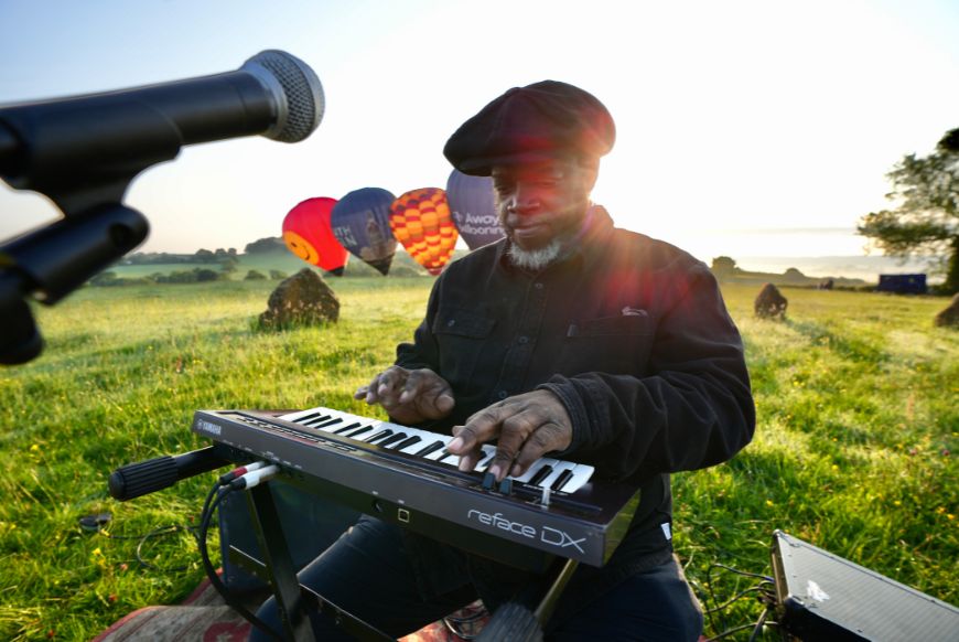 a man playing a keyboard in a field with hot air balloons behind